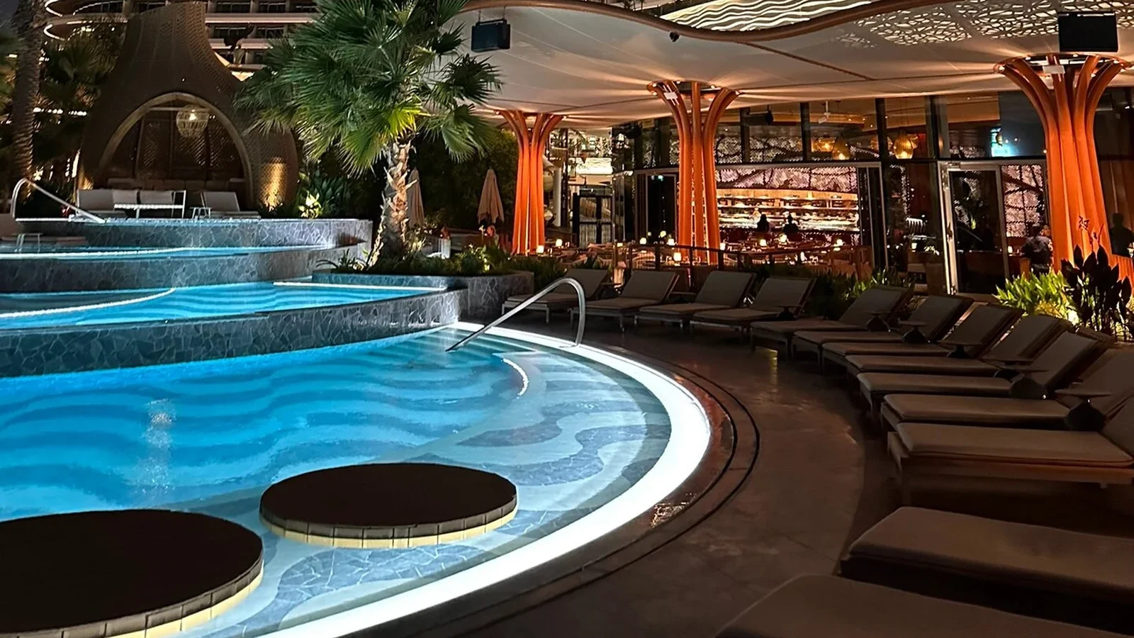 Cushioned resort pool loungers beside an illuminated swimming pool at night
